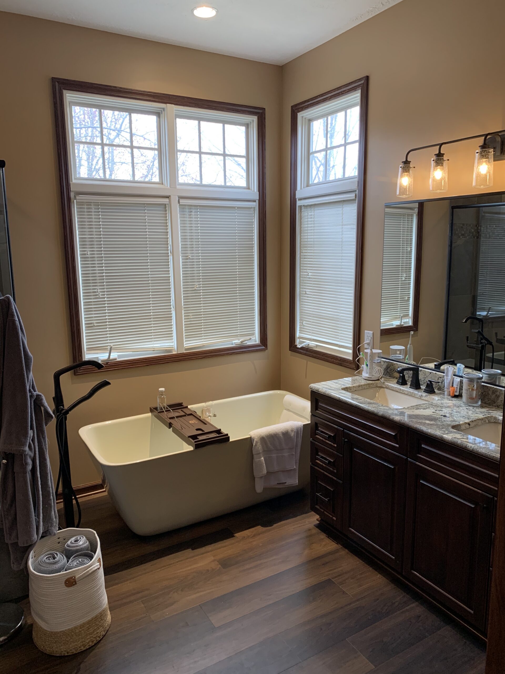 Classic bathroom, white soaking tub, double sink with dark cabinet storage, brown floor, windows