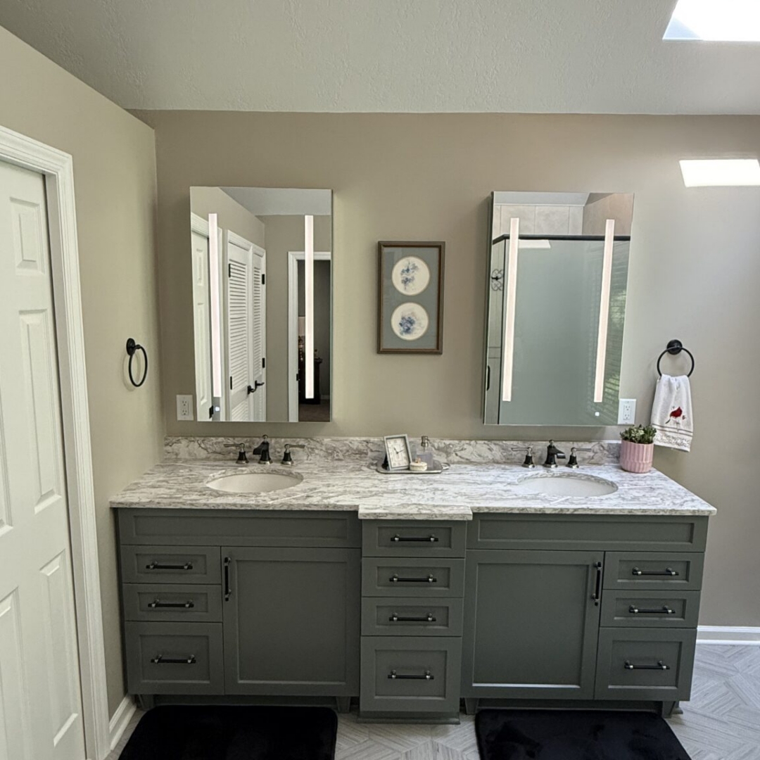 Renovated bathroom with vaulted ceiling, skylight, sage green double vanity with marble countertop, hexagon tile floor, arched window, and glass-enclosed shower with decorative tile border and built-in bench