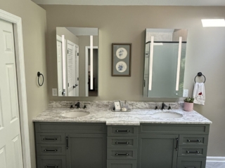 Renovated bathroom with vaulted ceiling, skylight, sage green double vanity with marble countertop, hexagon tile floor, arched window, and glass-enclosed shower with decorative tile border and built-in bench