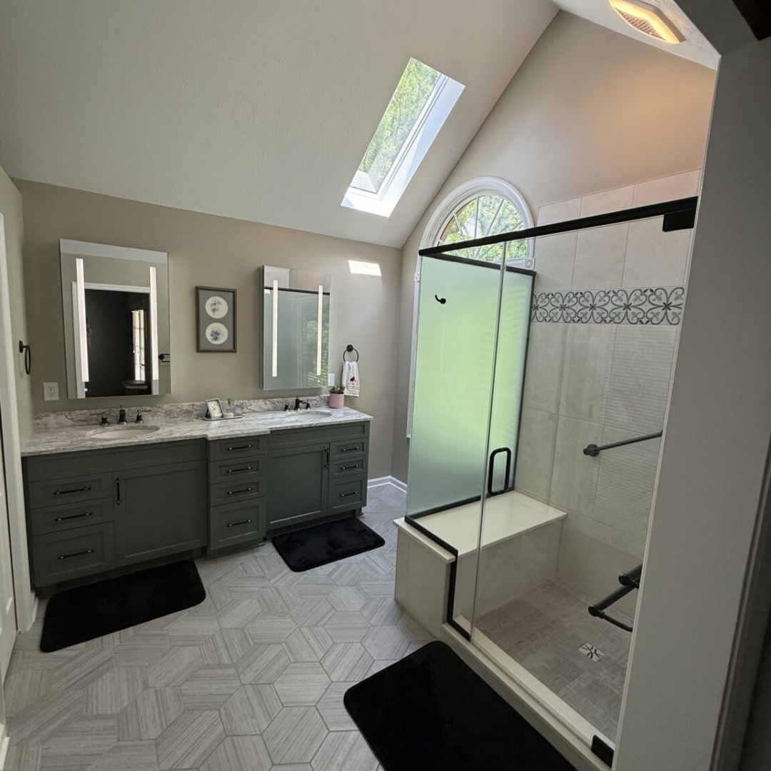 Renovated bathroom with vaulted ceiling, skylight, sage green double vanity with marble countertop, hexagon tile floor, arched window, and glass-enclosed shower with decorative tile border and built-in bench