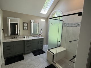 Renovated bathroom with vaulted ceiling, skylight, sage green double vanity with marble countertop, hexagon tile floor, arched window, and glass-enclosed shower with decorative tile border and built-in bench