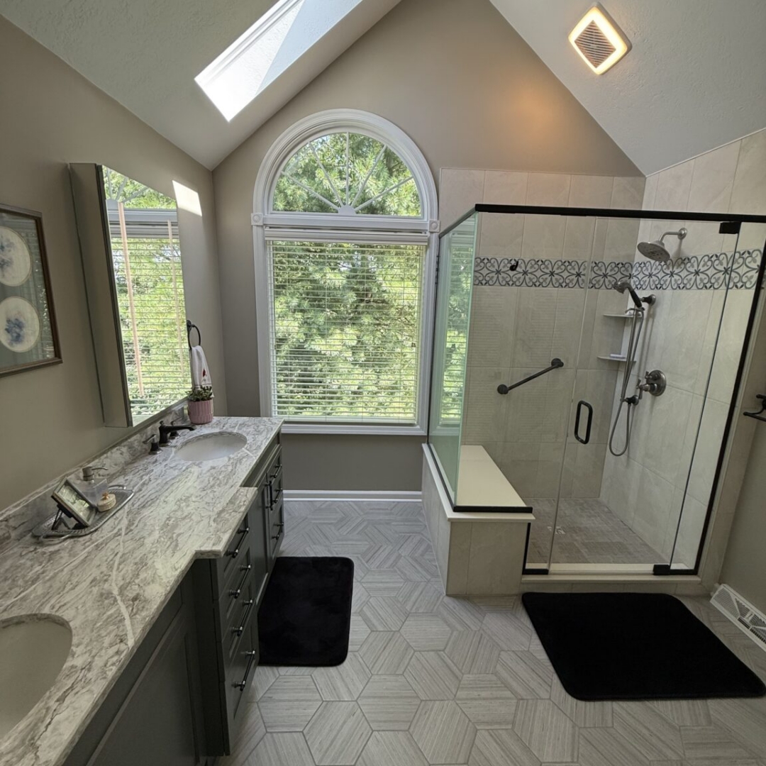 Renovated bathroom with vaulted ceiling, skylight, sage green double vanity with marble countertop, hexagon tile floor, arched window, and glass-enclosed shower with decorative tile border and built-in bench