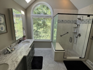 Renovated bathroom with vaulted ceiling, skylight, sage green double vanity with marble countertop, hexagon tile floor, arched window, and glass-enclosed shower with decorative tile border and built-in bench