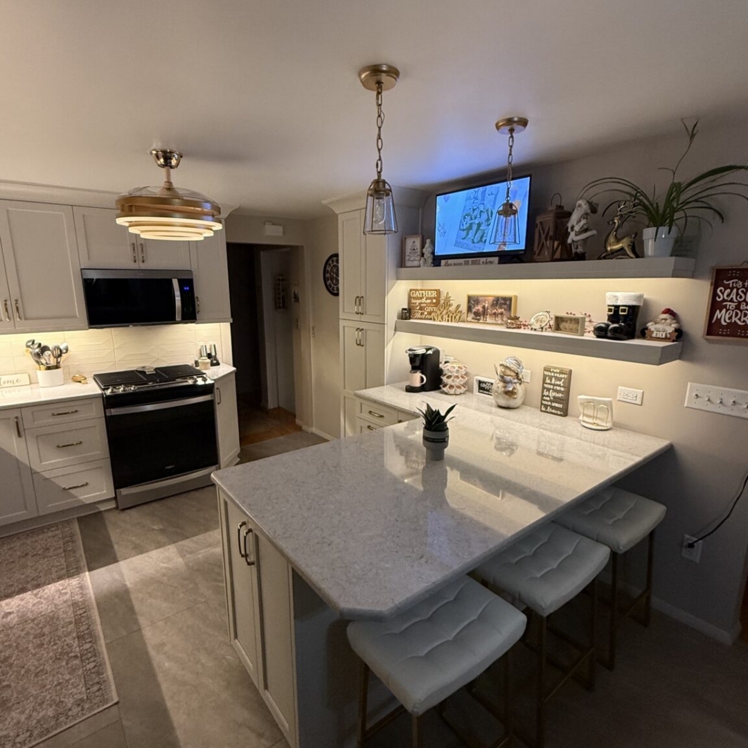 Kitchen remodel with white shaker cabinetry, quartz countertops, white subway tile backsplash, brass and gold hardware, retractable ceiling fan light fixture, glass pendant lights, white and gold bar stools, floating shelves with underlighting, and stainless steel appliances