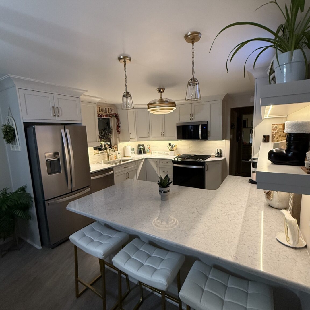 Kitchen remodel with white shaker cabinetry, quartz countertops, white subway tile backsplash, brass and gold hardware, retractable ceiling fan light fixture, glass pendant lights, white and gold bar stools, floating shelves with underlighting, and stainless steel appliances