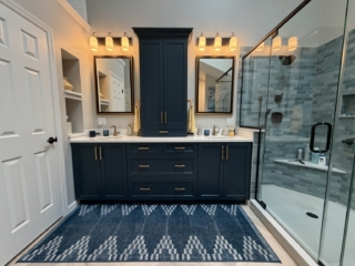 Luxury bathroom remodel featuring navy blue double vanity with brass hardware, quartz countertop, blue-grey subway tile walk-in shower, freestanding soaking tub with floor-mount tub filler, herringbone tile floor, and skylights