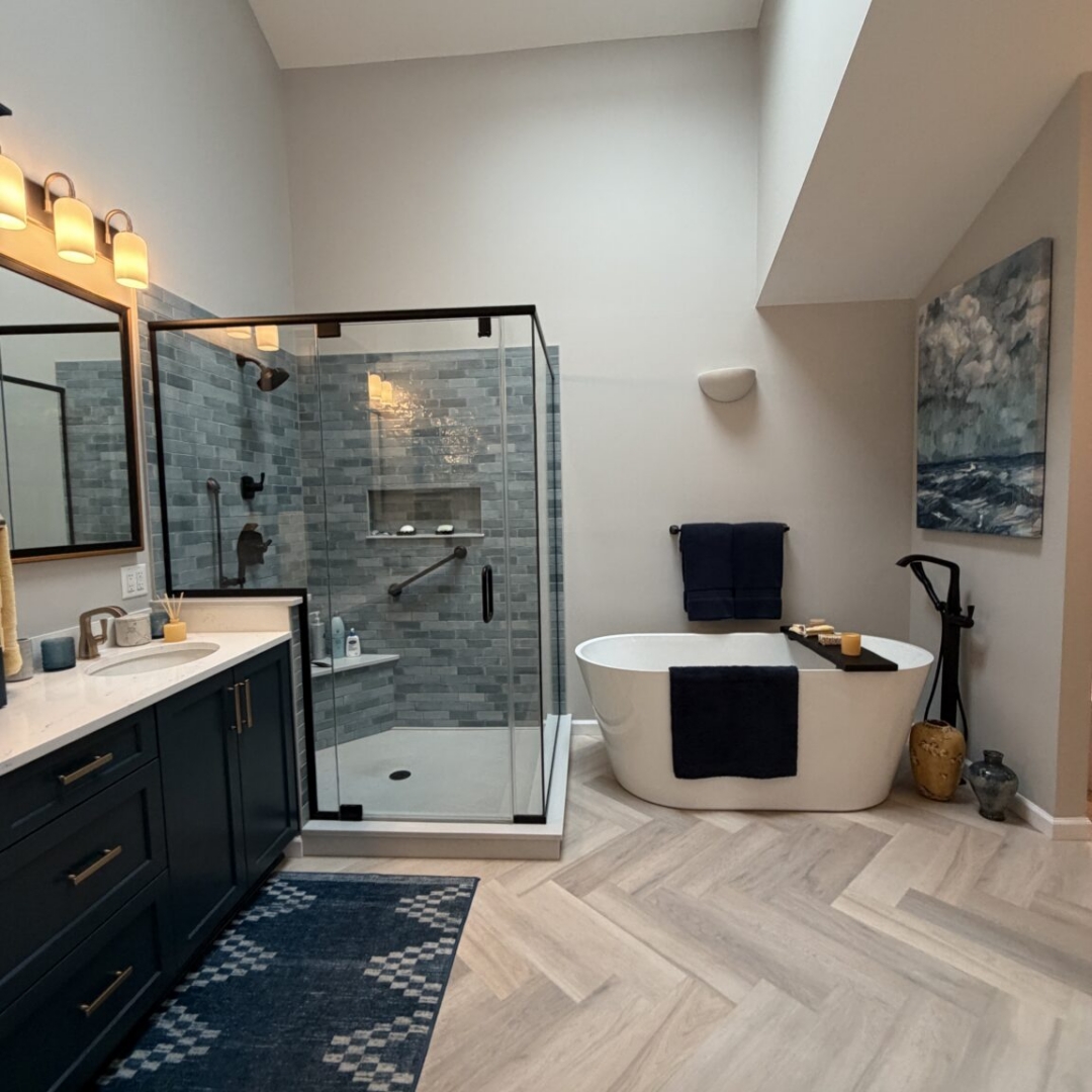 Luxury bathroom remodel featuring navy blue double vanity with brass hardware, quartz countertop, blue-grey subway tile walk-in shower, freestanding soaking tub with floor-mount tub filler, herringbone tile floor, and skylights