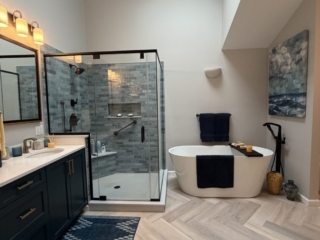 Luxury bathroom remodel featuring navy blue double vanity with brass hardware, quartz countertop, blue-grey subway tile walk-in shower, freestanding soaking tub with floor-mount tub filler, herringbone tile floor, and skylights