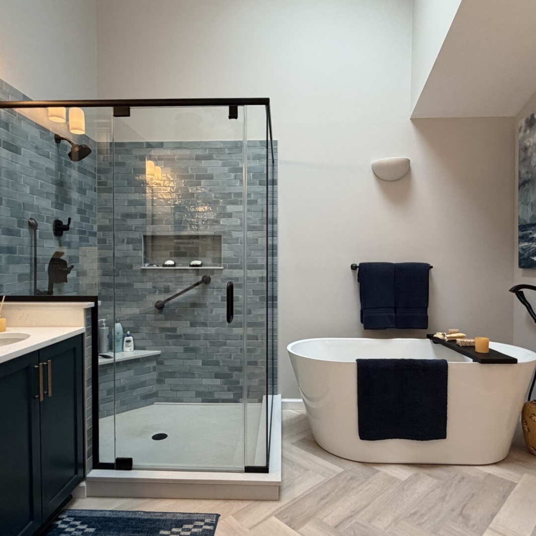 Luxury bathroom remodel featuring navy blue double vanity with brass hardware, quartz countertop, blue-grey subway tile walk-in shower, freestanding soaking tub with floor-mount tub filler, herringbone tile floor, and skylights