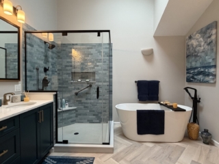 Luxury bathroom remodel featuring navy blue double vanity with brass hardware, quartz countertop, blue-grey subway tile walk-in shower, freestanding soaking tub with floor-mount tub filler, herringbone tile floor, and skylights