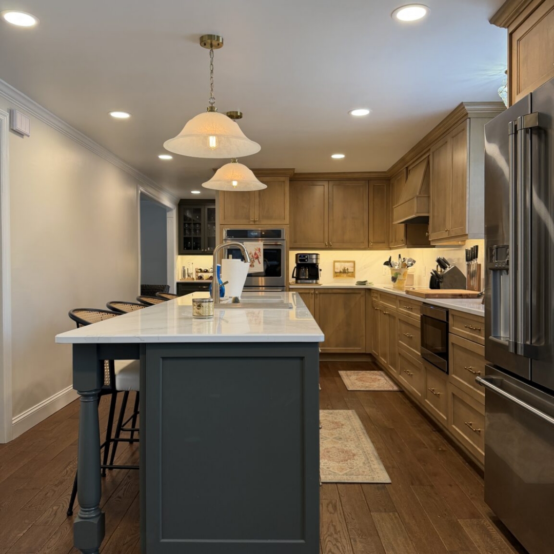 Kitchen remodel with warm taupe stained cabinetry, contrasting slate blue island with brass hardware, quartz countertops, marble-look tile backsplash, custom range hood, hardwood floors, pendant lighting, and stainless steel appliances